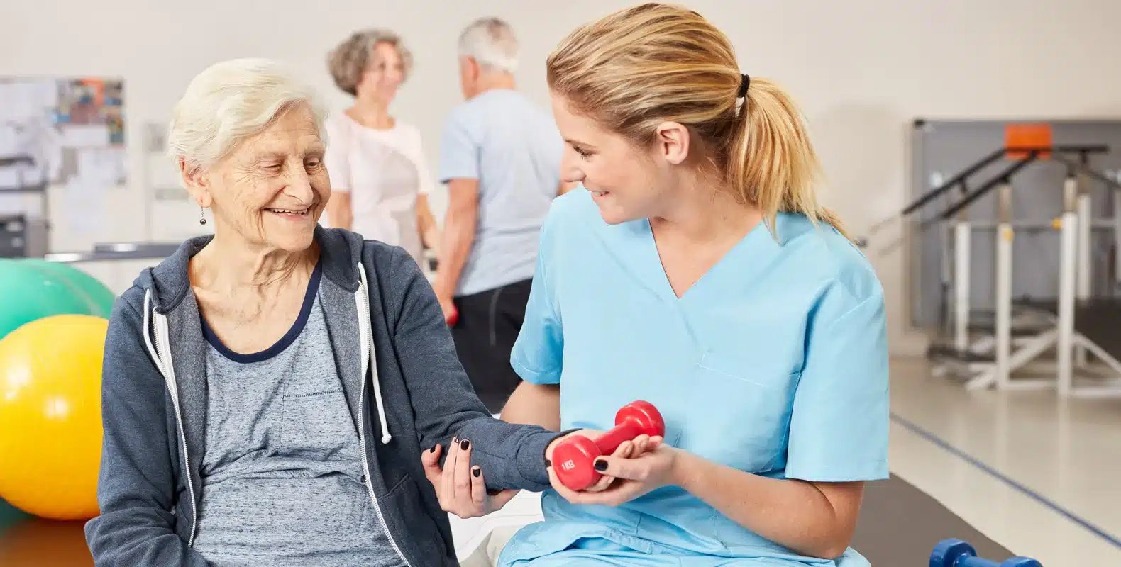 Elderly woman and caregiver smiling while exercising with red dumbbells in a bright, well-equipped rehabilitation center.