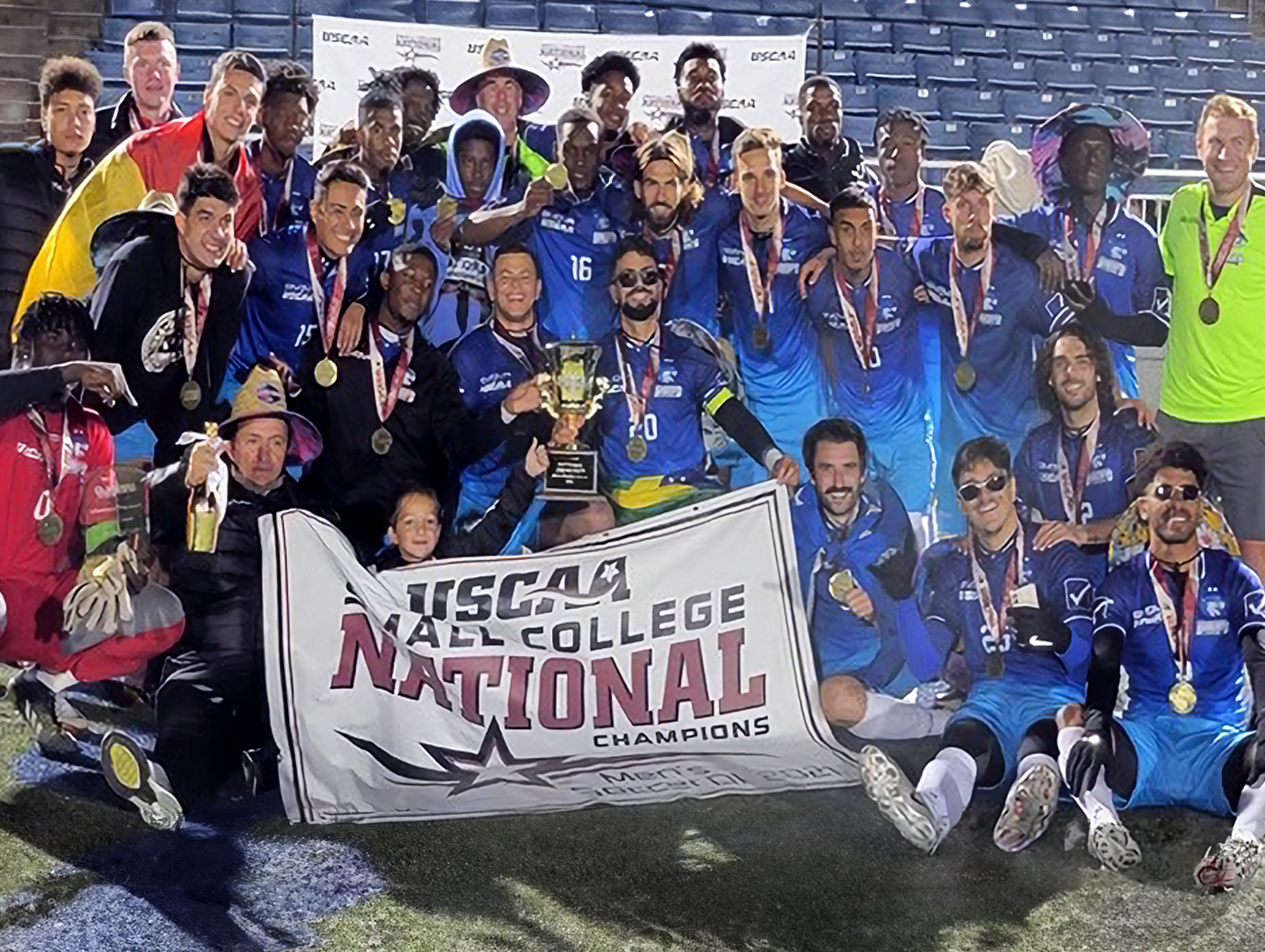 A college soccer team celebrates winning the national championship, holding medals, a trophy, and a championship banner.