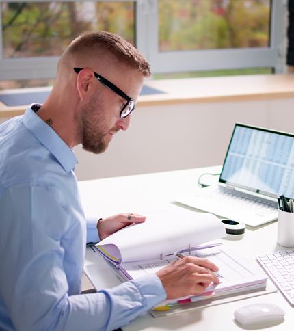 A man with glasses sits at a desk, reading and writing in a notebook. A laptop with medical coding charts on the screen and office supplies are on the desk. Large windows are in the background.