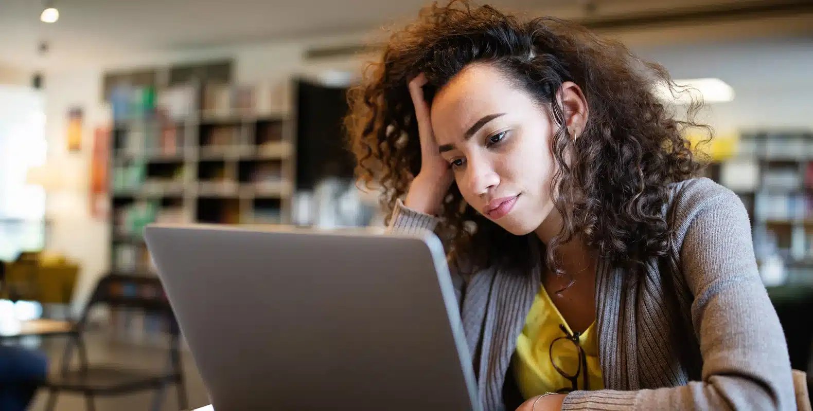 Person with curly hair sitting at a desk, looking at a laptop with a pensive expression, background of bookshelves.