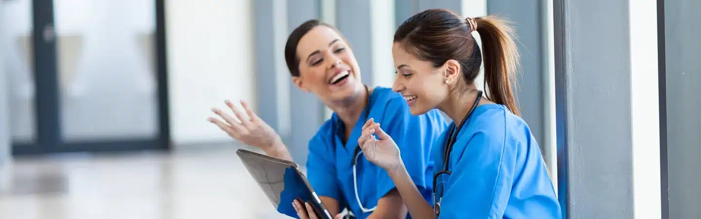 Two healthcare professionals in blue scrubs smiling and looking at a tablet near a large window.