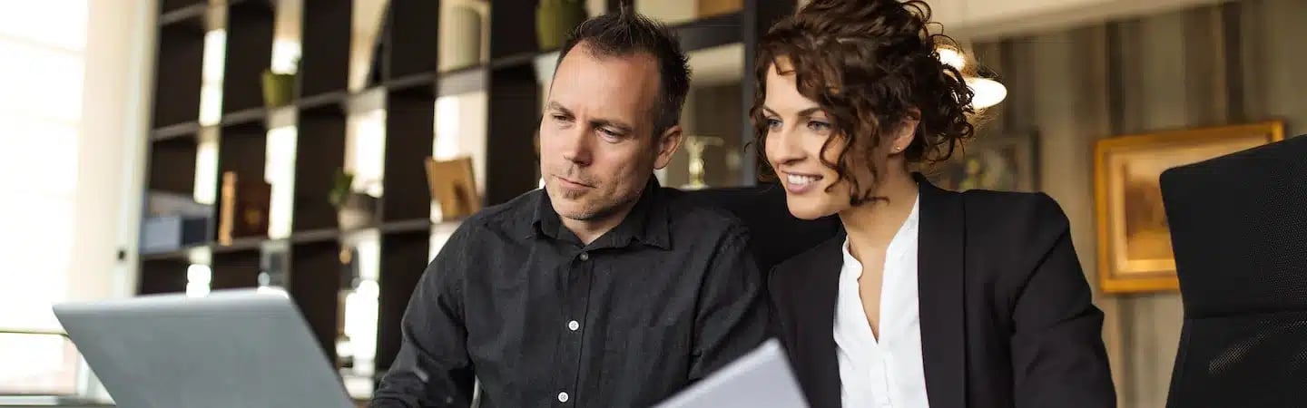 Two people in a modern office setting, working together on a laptop. The background shows shelves with various items.