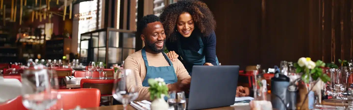 Two restaurant staff members happily looking at a laptop screen while sitting inside the restaurant with set tables around them.