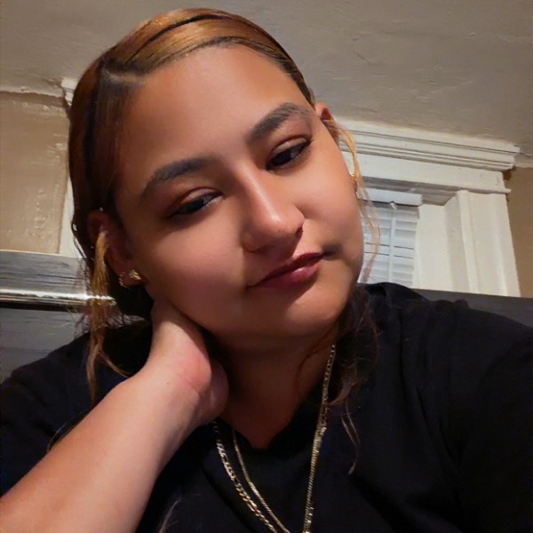 A woman with honey-toned hair resting her chin on her hand, wearing a black shirt and a gold chain.