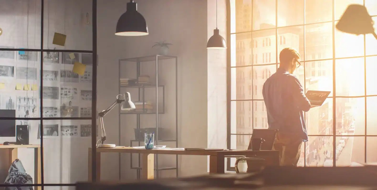 A person stands near a large window in a sunlit office holding a laptop, with desks and shelves in the background.