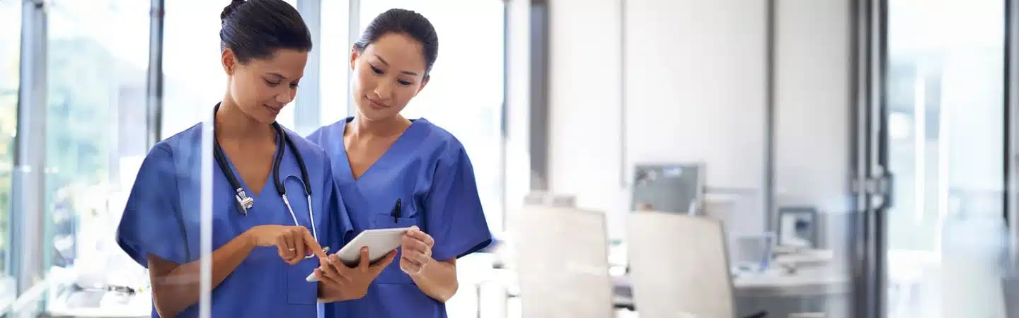 Two healthcare professionals in blue scrubs discussing information on a tablet in a bright office setting.