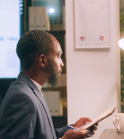A payroll clerk in a suit sits at a desk holding a clipboard and pen, looking thoughtfully at a wall-mounted chart with graphs, next to a lamp and coffee mug. Office shelves and a large screen complete the professional setting.