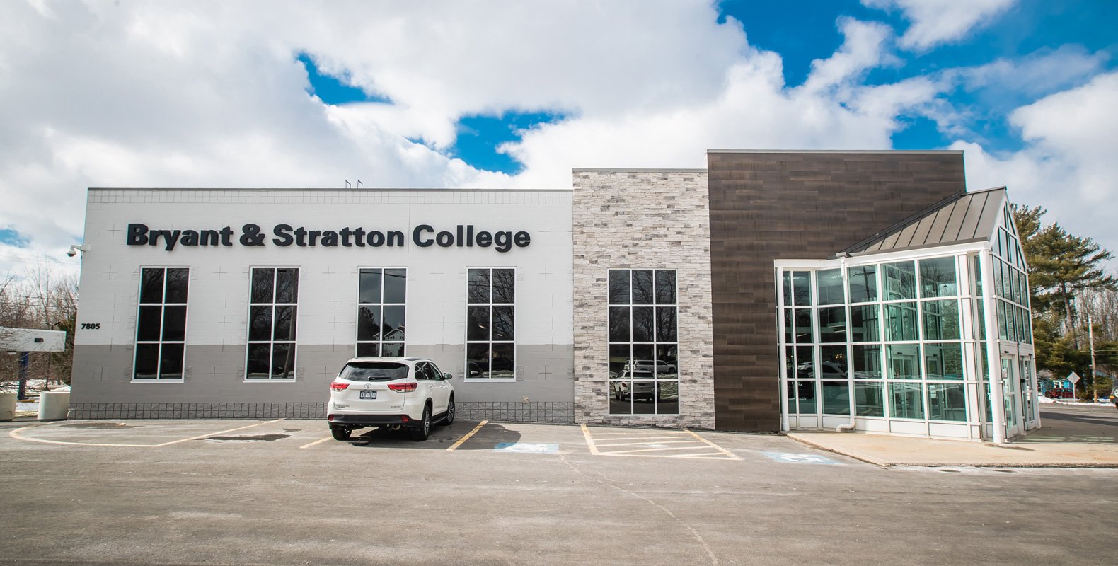 A modern building with large windows and a sign reading "Bryant & Stratton College" on the exterior in Liverpool, NY, with a white SUV parked in front under a partly cloudy sky.