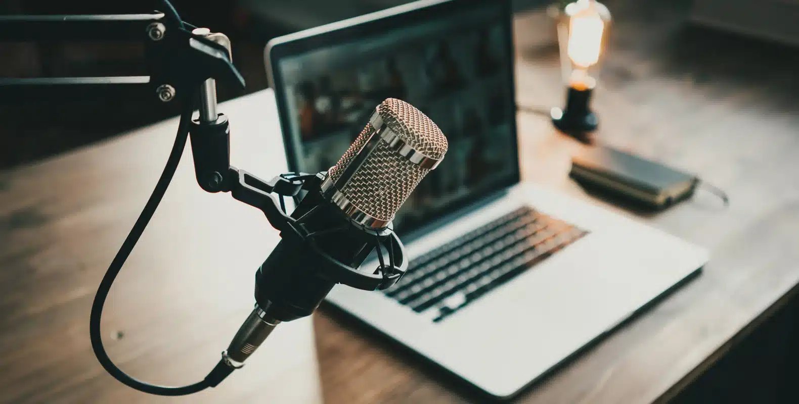 Close-up of a microphone on a stand in front of a laptop on a wooden desk, with a notebook and a light bulb in the background.