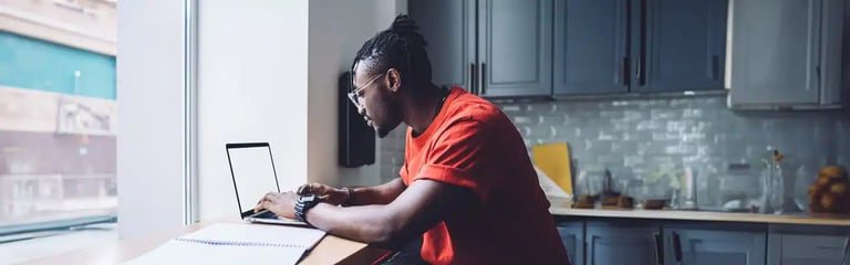 Person in red shirt using laptop at a kitchen counter near a window, with a notebook and glassware in the background.