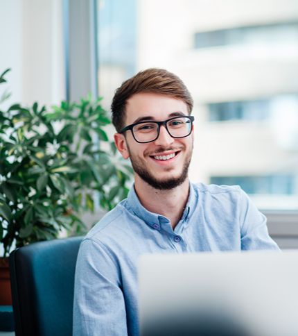 A young administrator with glasses and a light blue shirt smiles while sitting at a desk with a laptop. There is a green plant and a large window with blurred buildings in the background.