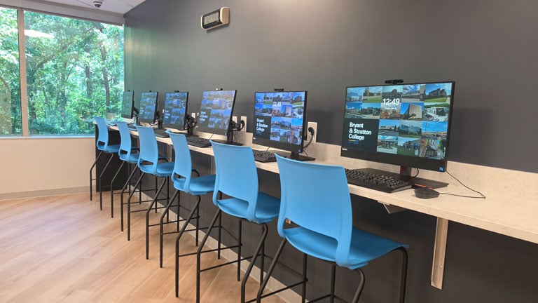 A row of desktop computers with blue chairs in a modern computer lab at a Richmond college, featuring large windows with views of green trees. The screens display the Bryant & Stratton College login page, highlighting colleges in Richmond VA.