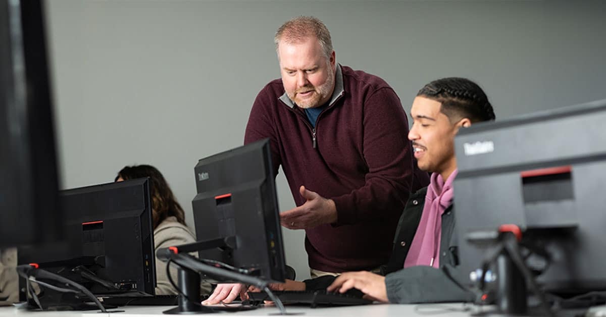 Instructor assisting a student at a computer lab while other students work at their stations in the background.