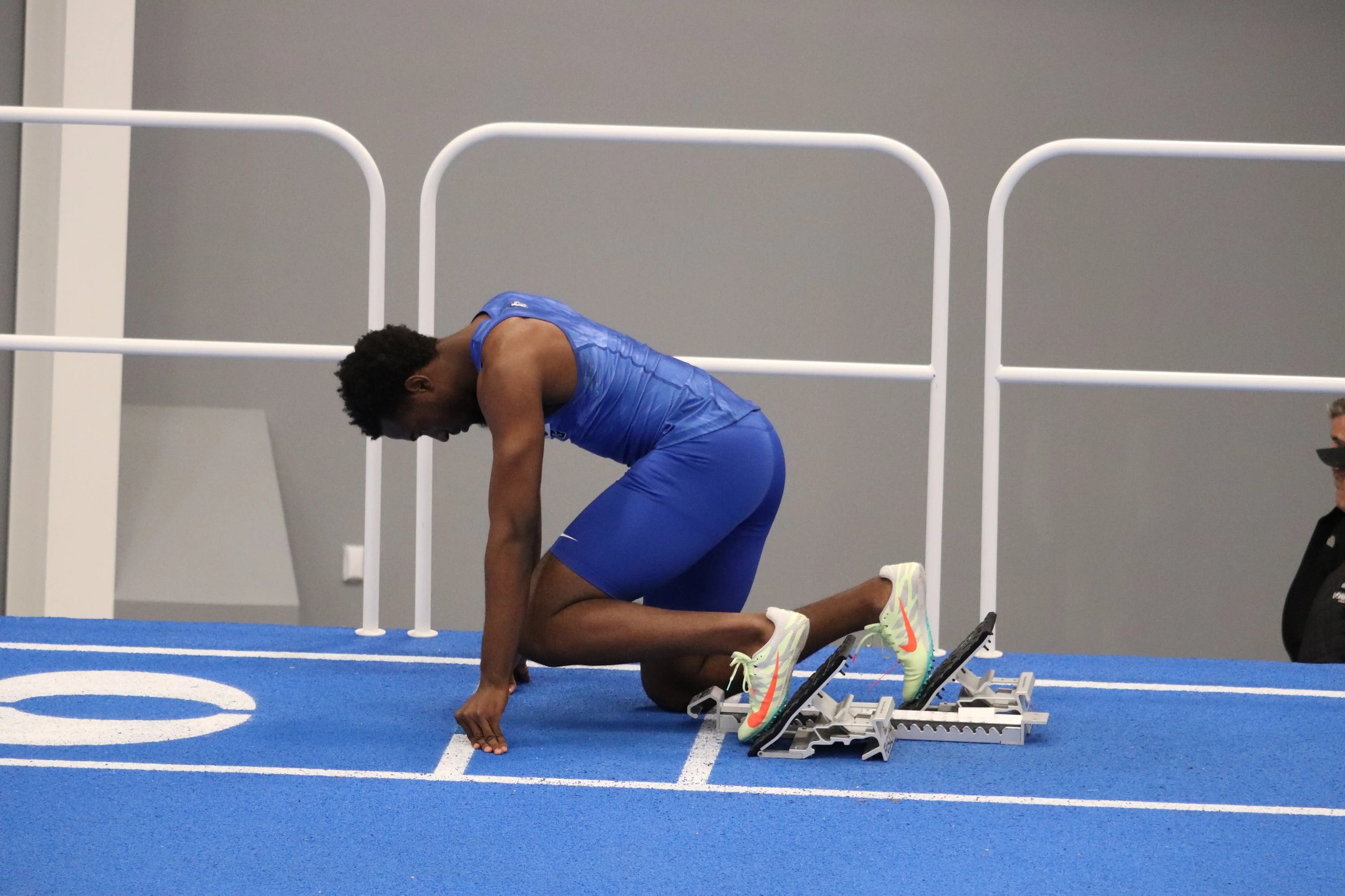 Athlete in blue prepares for a sprint on an indoor track, one knee down and hands positioned at the starting line.