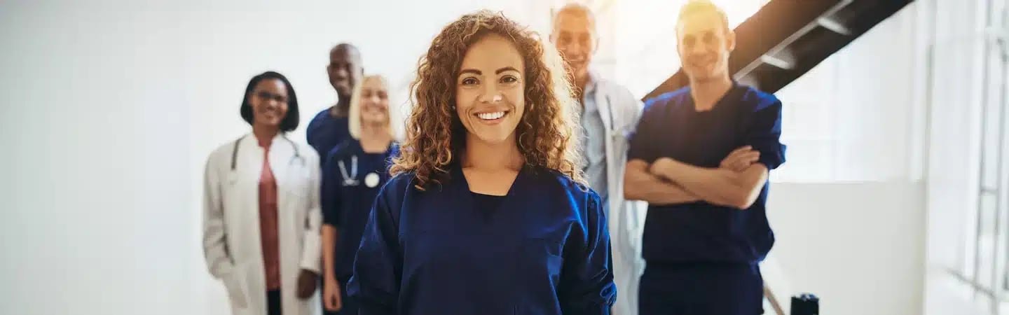 A group of medical professionals smiling, with a woman in blue scrubs in the front and others standing behind her.