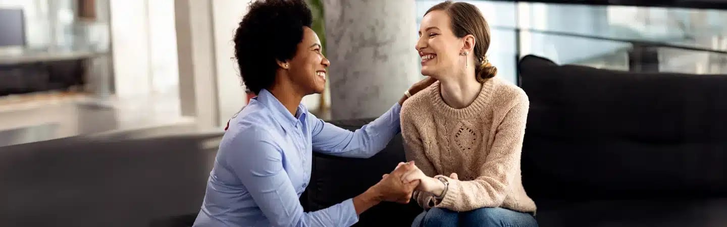 Two women smiling and holding hands while sitting on a couch, engaged in a friendly and supportive conversation.