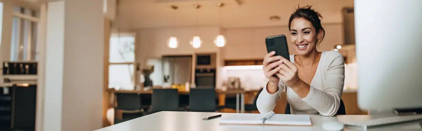 Woman smiling while looking at her phone, seated at a desk with a notebook, in a bright kitchen.