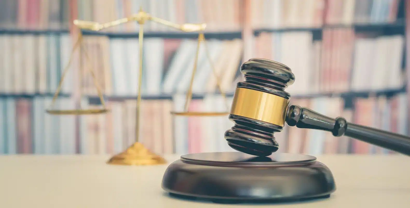 A judge's gavel and sound block sit on a desk, with a scales of justice and rows of books blurred in the background.