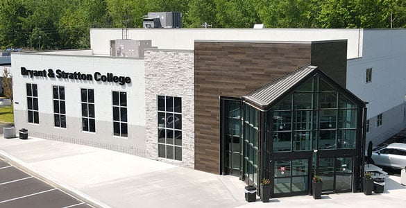 Modern building with large windows and a glass entrance displaying the sign "Bryant & Stratton College." Located in Liverpool, NY, the exterior features white, stone, and wood-like panels, surrounded by greenery and a parking lot.