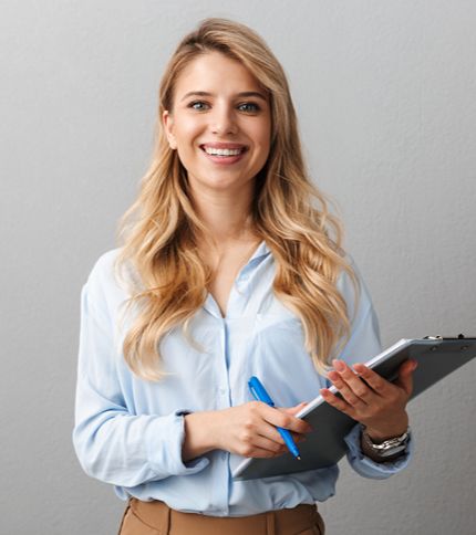 A smiling woman with long blonde hair, wearing a light blue shirt, holds a clipboard and blue pen—ready to tackle admin assistant job duties—standing in front of a plain gray wall.