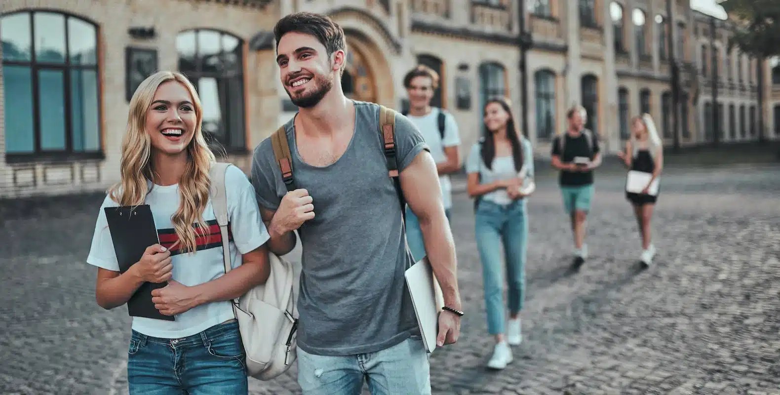 Group of smiling young adults walking on a cobblestone street, carrying backpacks and books near an old building.