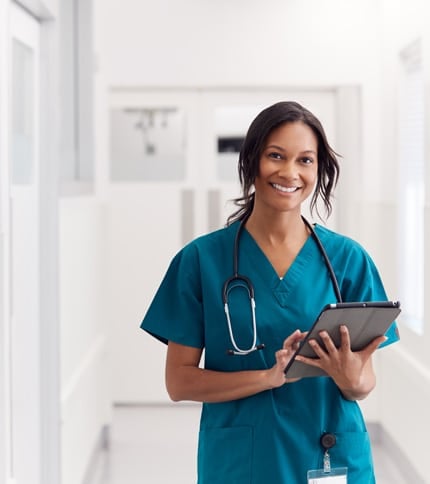 A Registered Nurse in teal scrubs with a stethoscope around their neck smiles while using a tablet in a bright hospital corridor.