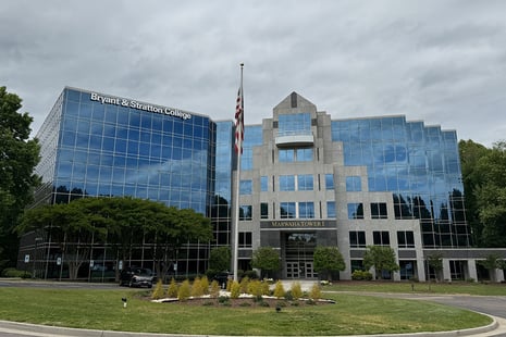 A modern glass office building, home to Bryant & Stratton College—one of the leading colleges in Richmond, VA—displays “Marathon Tower I” above the entrance. An American flag stands in front, surrounded by trees.