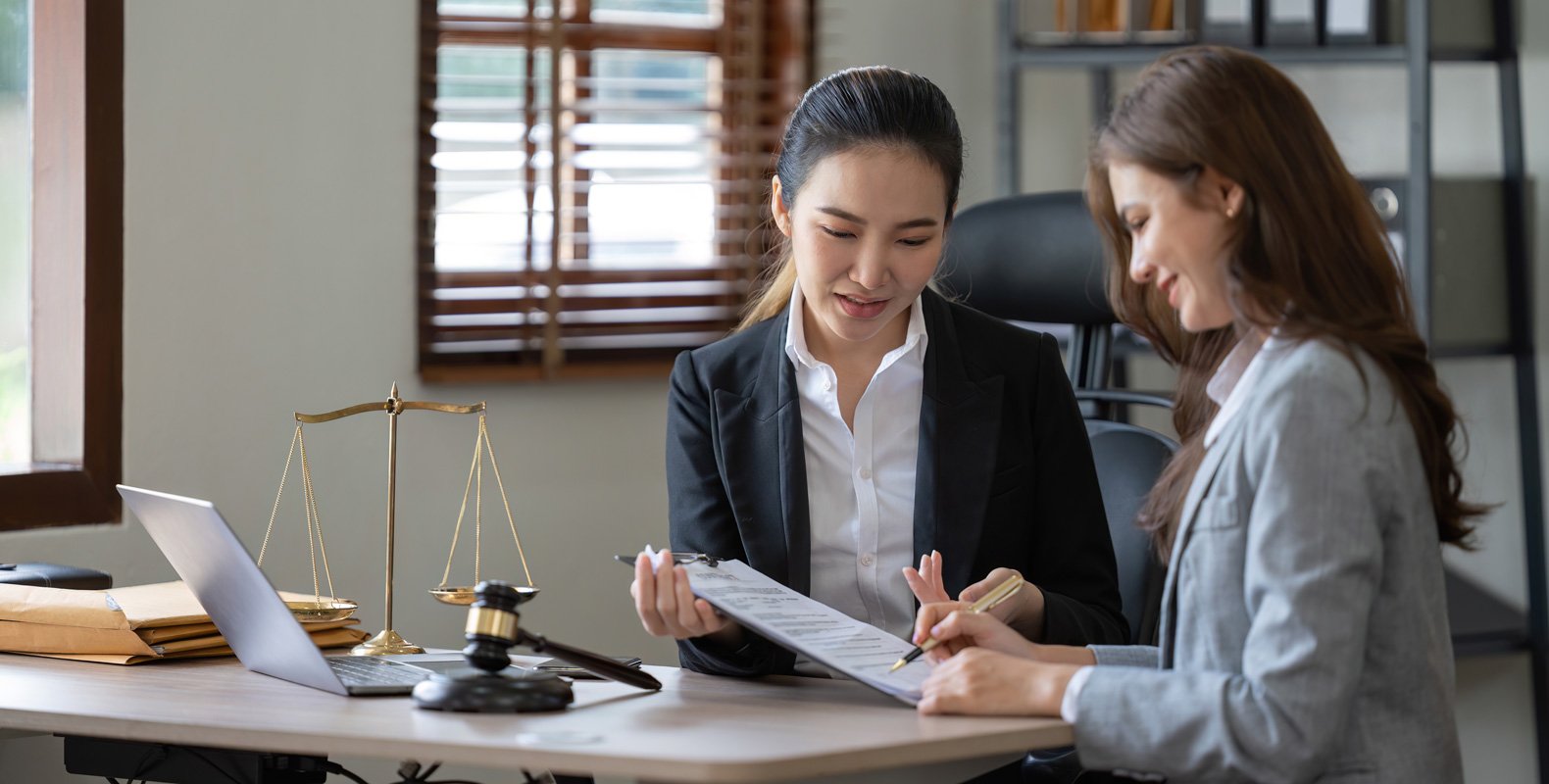 Two women in business attire sit at a desk discussing a document. Legal scales, a gavel, a laptop, and folders are on the desk, suggesting a professional legal or business consultation.