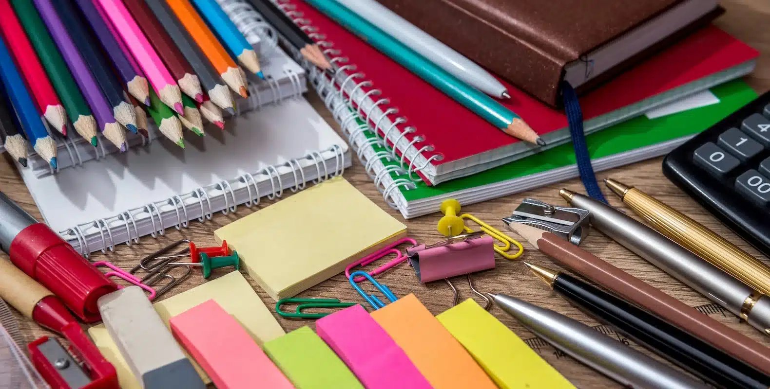 A variety of colorful office supplies, including pens, pencils, sticky notes, notebooks, and clips, spread out on a table.