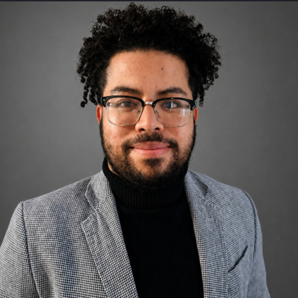 A man wearing glasses, a black turtleneck, and a patterned blazer poses against a gray background.