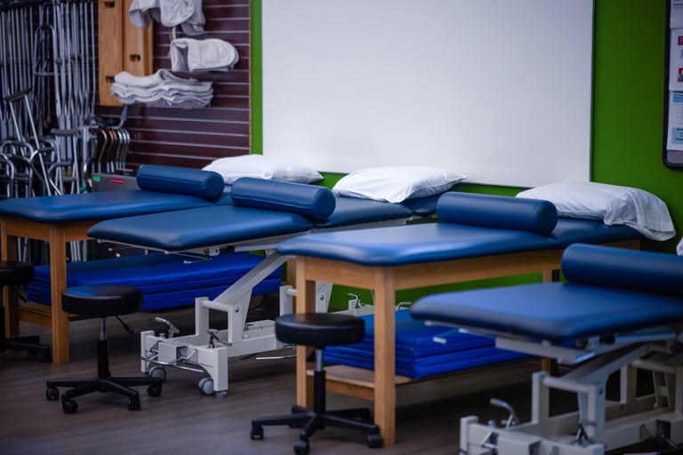 A row of blue padded physical therapy tables with pillows, stools, and folded towels in a medical clinic in Orchard Park NY, with mobility aids visible in the background.
