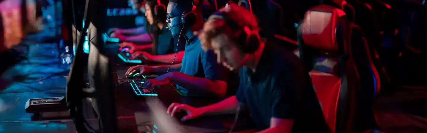A row of gamers wearing headsets intensely focused on their screens in a dimly lit gaming arena with blue and red lighting.