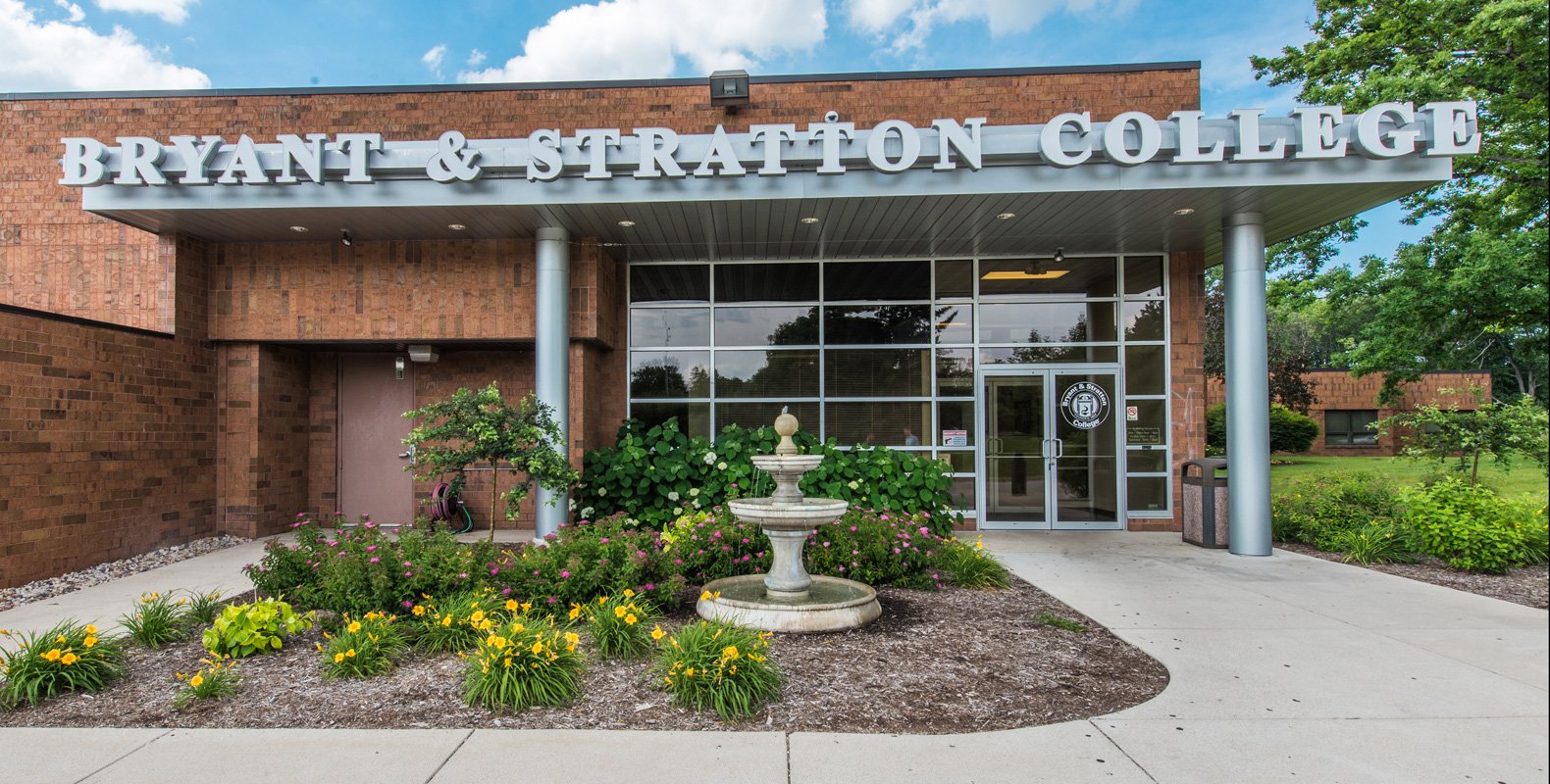 The entrance of Bryant & Stratton College in Wauwatosa, WI features large glass windows, brick walls, a double-door entrance, a decorative fountain, and vibrant flower beds. The college name is prominently displayed above the entrance.