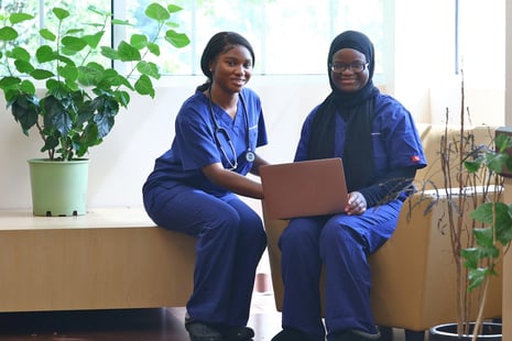 Two medical professionals in blue scrubs smiling and using a laptop in a bright room with green plants.