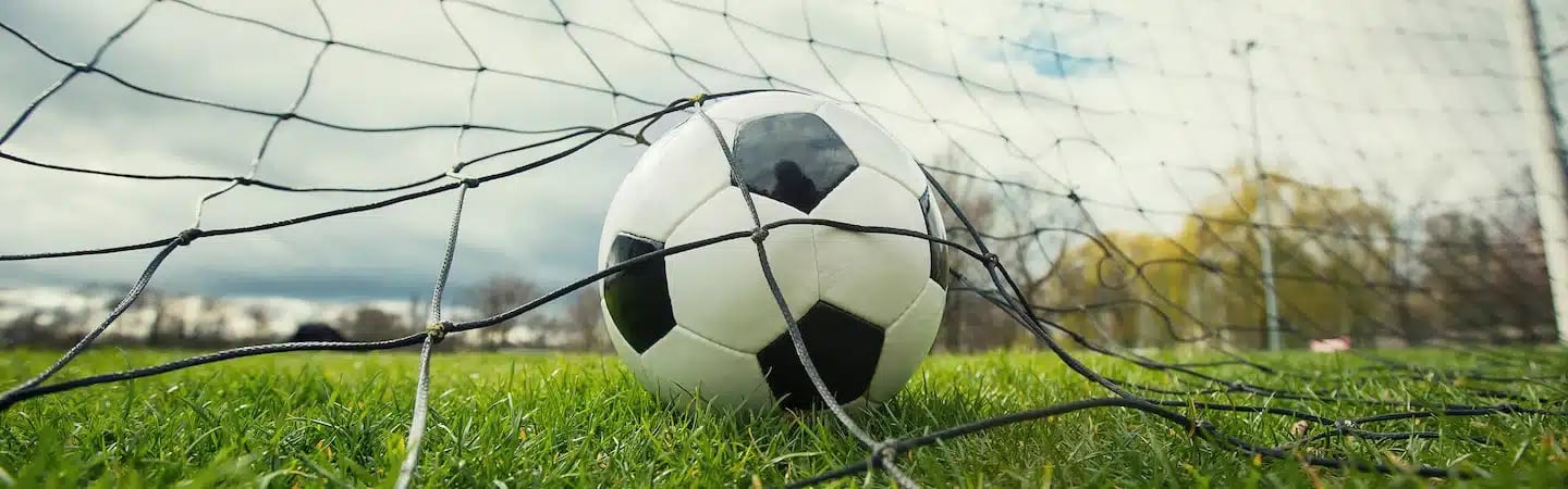 Close-up of a soccer ball tangled in the net of a goal on a grassy field with a cloudy sky in the background.
