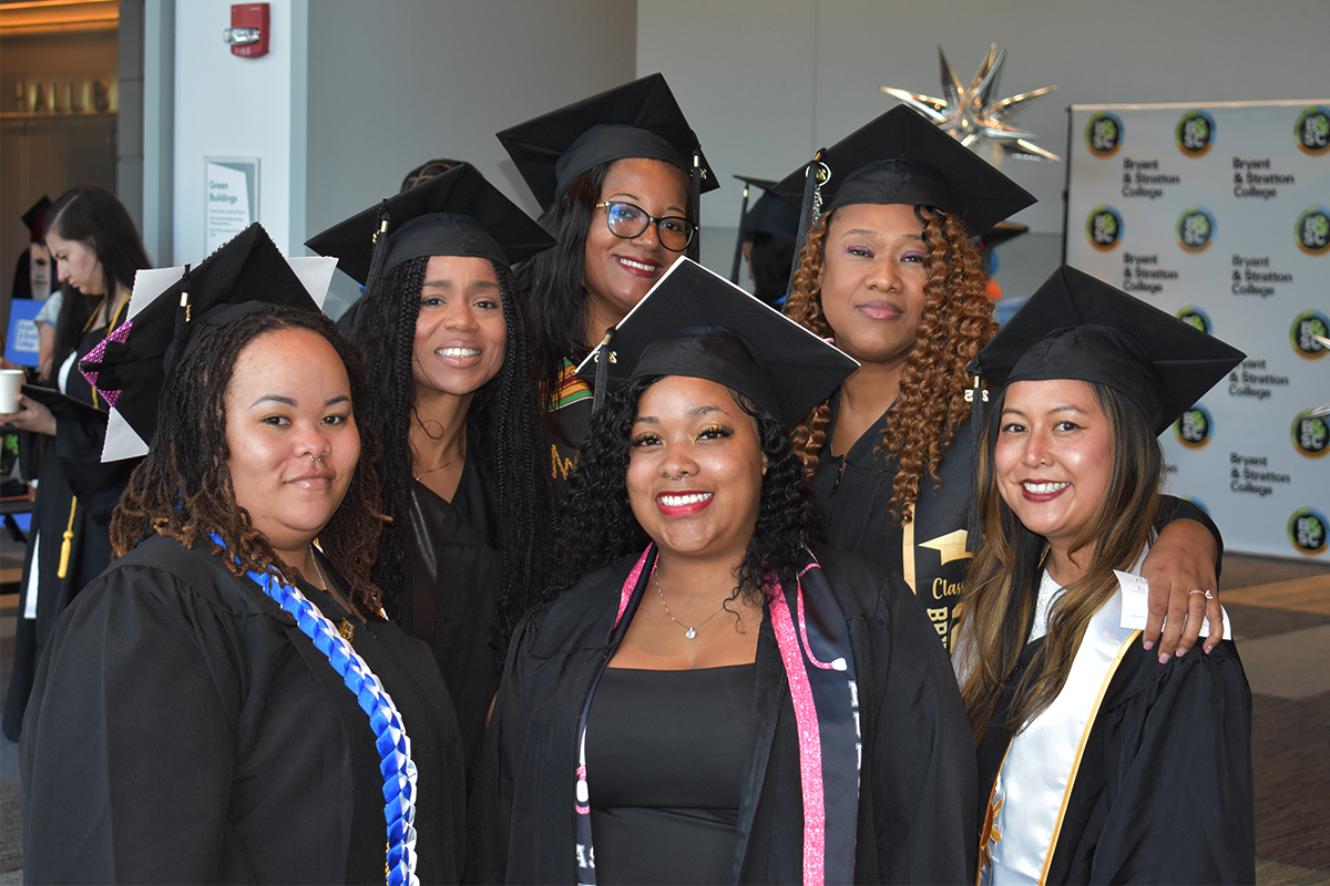 Six women in graduation caps and gowns stand together smiling indoors, with a background featuring a Broward College sign and other graduates.