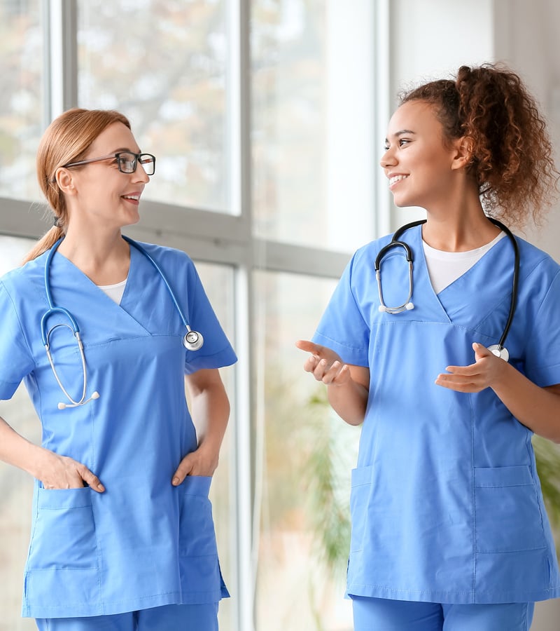 Two healthcare professionals in blue scrubs, each wearing a stethoscope, stand indoors smiling and talking to each other in front of large windows.