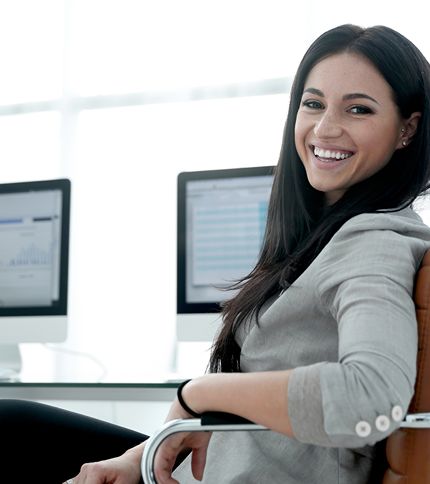 A Marketing Manager with long dark hair, wearing a light gray blazer, smiles while sitting in an office chair. Two computer monitors displaying charts and data are visible in the background.