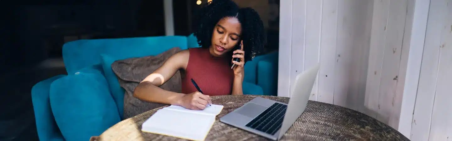 A woman sits at a table with a laptop, writing in a notebook while talking on the phone.