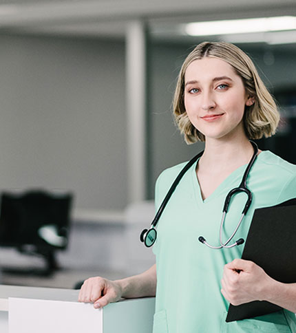 A healthcare professional in green scrubs, representing the excellence of nursing programs in Rochester, NY, stands confidently holding a black folder. The backdrop is a bustling medical office with a desk and computer.