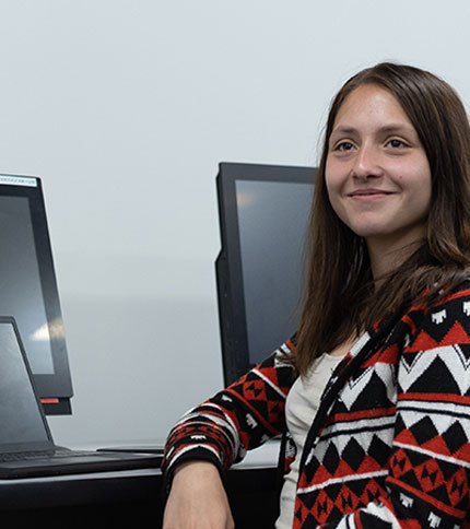 A person with long hair smiles while sitting in front of computer screens, perhaps pursuing an online digital marketing degree. They're wearing a patterned sweater in a room with neutral-colored walls.