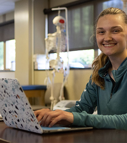 A person in a teal jacket is smiling and working on a laptop in a room with a skeleton model in the background. The room has large windows and appears to be well-lit.