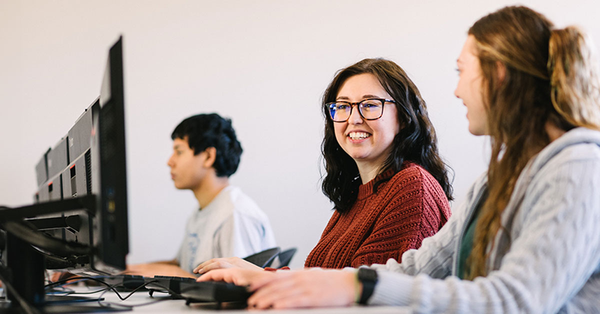 Three students are seated at a row of computers in a classroom. The student in the middle, wearing glasses and a red sweater, is smiling at the student on the right. The third student is focused on the screen.