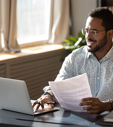 A man with glasses and a beard is sitting at a desk, smiling while holding paperwork and using a laptop. Sunlight filters through a window in the background.