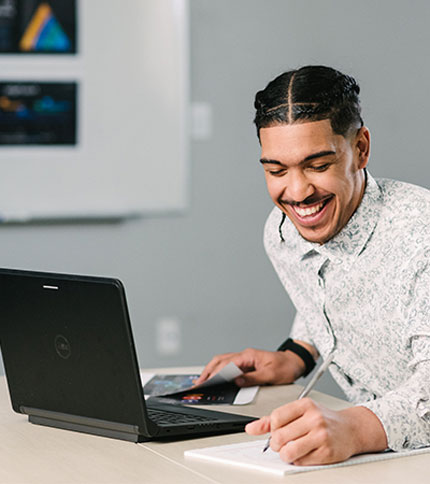 A man smiles while sitting at a desk with a laptop in front of him. He is writing notes on a notebook with a pen. A whiteboard with visible designs is in the background.