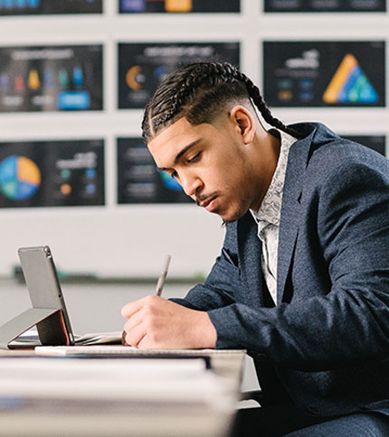 A person with braided hair in a suit is sitting at a desk, writing in a notebook. A tablet is propped up nearby. In the background, blurred charts and graphs are displayed on a whiteboard.