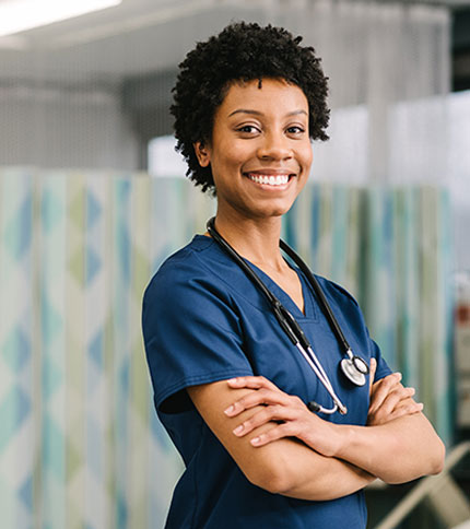 A smiling healthcare professional stands with arms crossed, wearing navy blue scrubs and a stethoscope around their neck. They are in a medical setting with a patterned privacy curtain in the background.