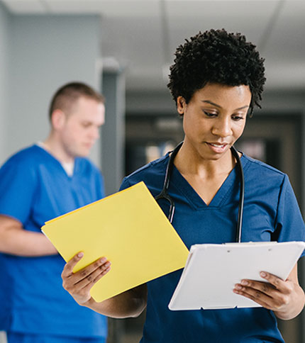 A healthcare professional wearing blue scrubs and a stethoscope reads a document and a tablet. In the background, another person in blue scrubs is blurred, standing in a hallway.