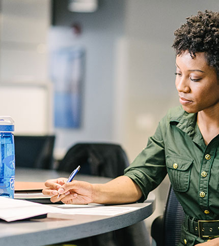 A person in a green shirt sits at a table, writing on a notepad with a pen. A blue water bottle and some papers are also on the table. The setting appears to be an office or meeting room.