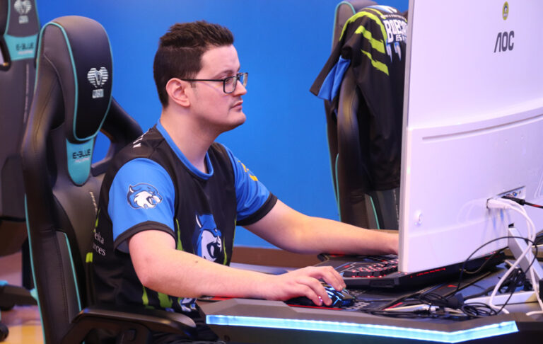 A man wearing glasses and a blue esports jersey focuses on playing a computer game with a large white monitor.